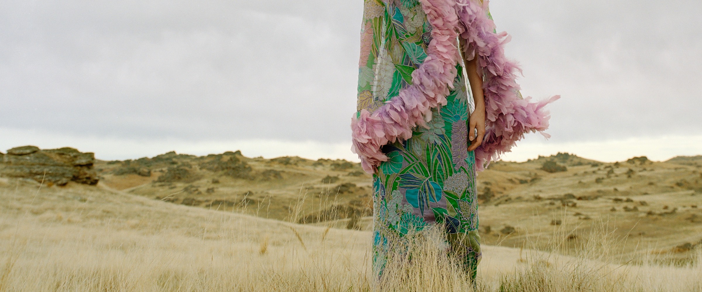 A model wearing a dress patterned in blue, pink and green, standing on a golden hills with a pink boa draped over her